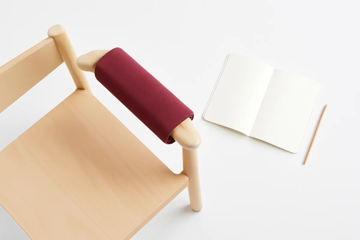 Vista cenital de una silla de madera clara con el manguito de tensión granate colocado sobre el reposabrazos, y al lado un cuaderno abierto en blanco con un lápiz; sin aparato, sin pantallas, sin texto